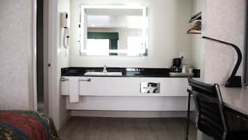 A compact hotel room with a white sink, a mirror above it, a black chair, and a bed with a colorful blanket.