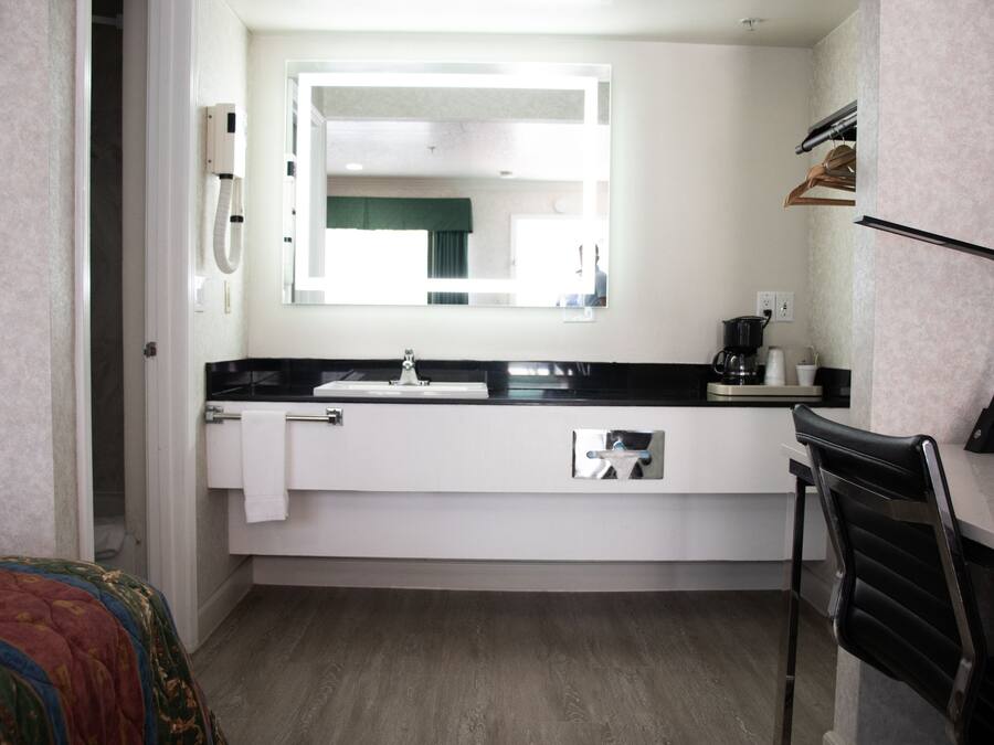 A compact hotel room with a white sink, a mirror above it, a black chair, and a bed with a colorful blanket.
