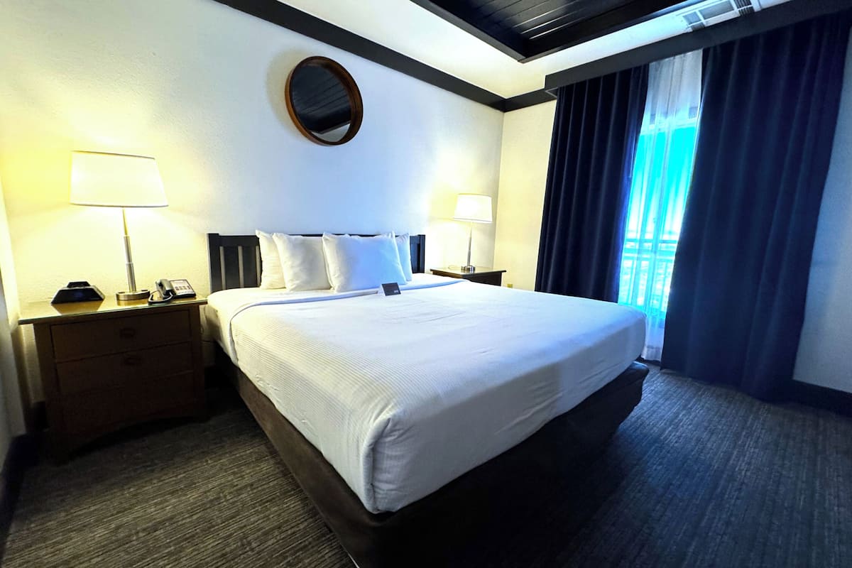 A hotel room with a large bed, two bedside tables, a round mirror, and a view of the outdoors through a window with curtains.