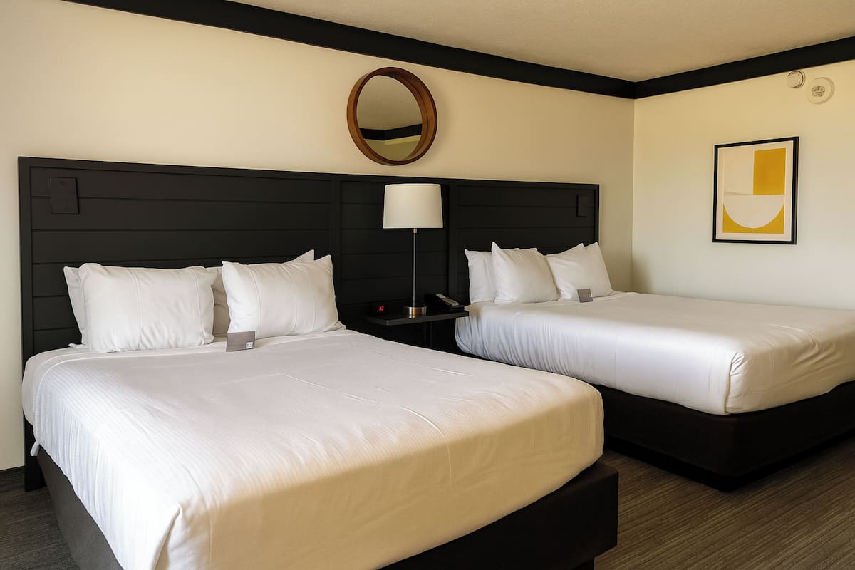 A hotel room with two beds, a bedside table with a lamp, a wall clock, and a framed abstract art piece.