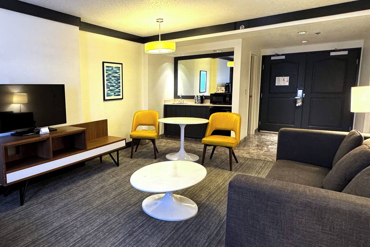 A hotel room with a TV, sofa, two yellow chairs, a small table, and a kitchen area with a microwave.