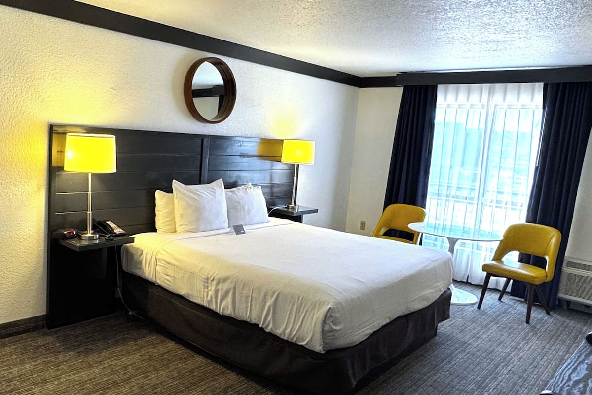 A hotel room with a large bed, two bedside tables with lamps, a round mirror, a small table with a chair, and a view of the city through the window.