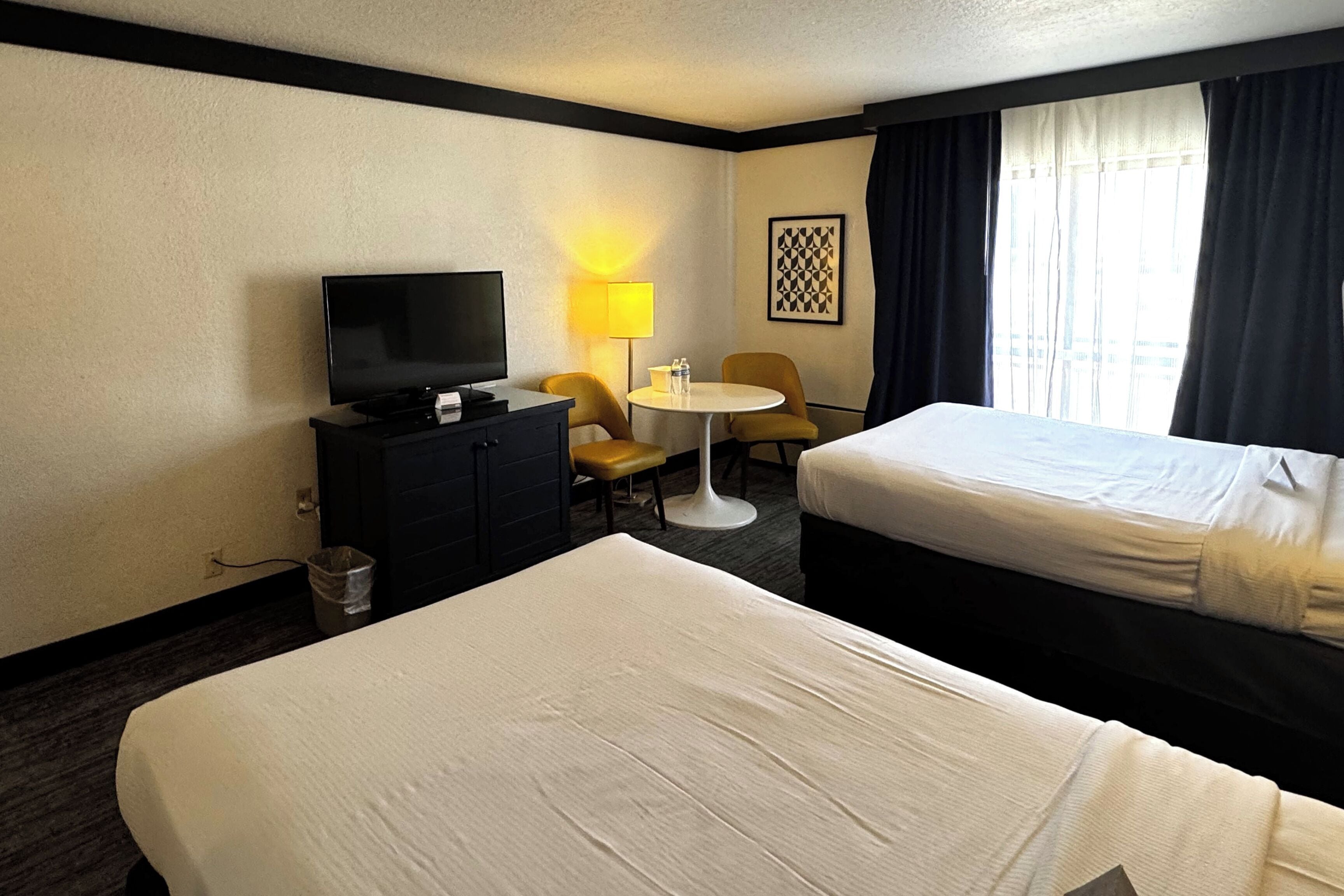 Une chambre d’hôtel avec deux lits, une télévision, un bureau avec une chaise, une lampe et une fenêtre avec des rideaux.