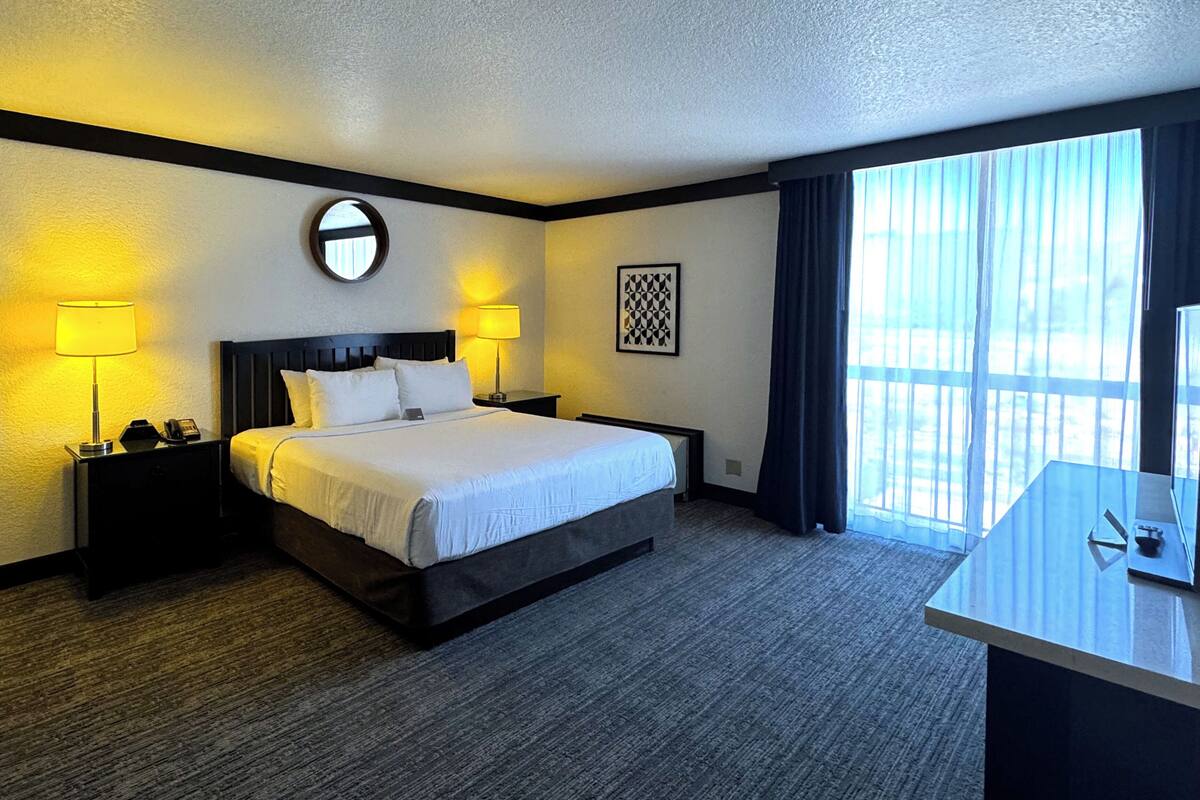 A hotel room with a large bed, two bedside lamps, a round mirror, a framed artwork, and a desk with a chair.