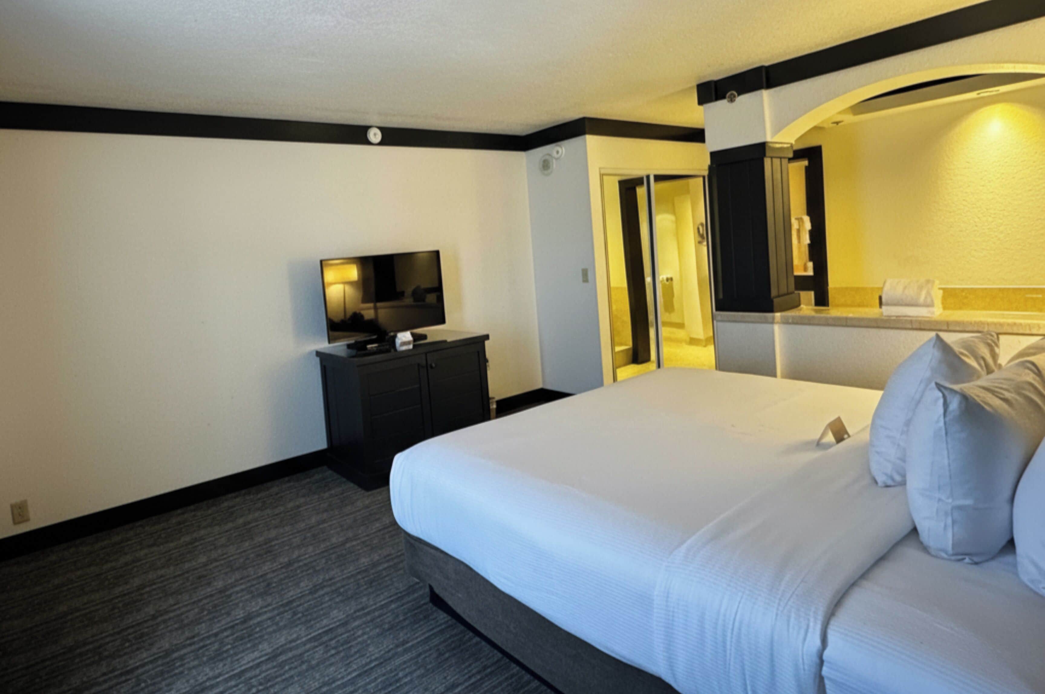 Une chambre d’hôtel avec un grand lit, un téléviseur à écran plat posé sur une commode, un miroir et une salle de bain visible à travers une arche.