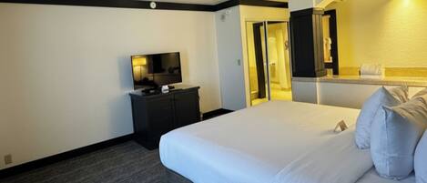 Une chambre d’hôtel avec un grand lit, un téléviseur à écran plat posé sur une commode, un miroir et une salle de bain visible à travers une arche.