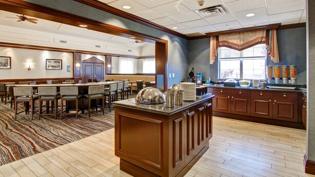 A spacious hotel lobby with a large dining area, a kitchen counter, and a seating area with tables and chairs.
