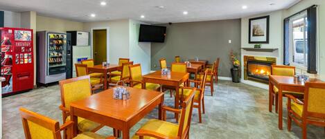 A hotel dining area with multiple wooden tables and chairs, a television mounted on the wall, a fireplace, and vending machines.