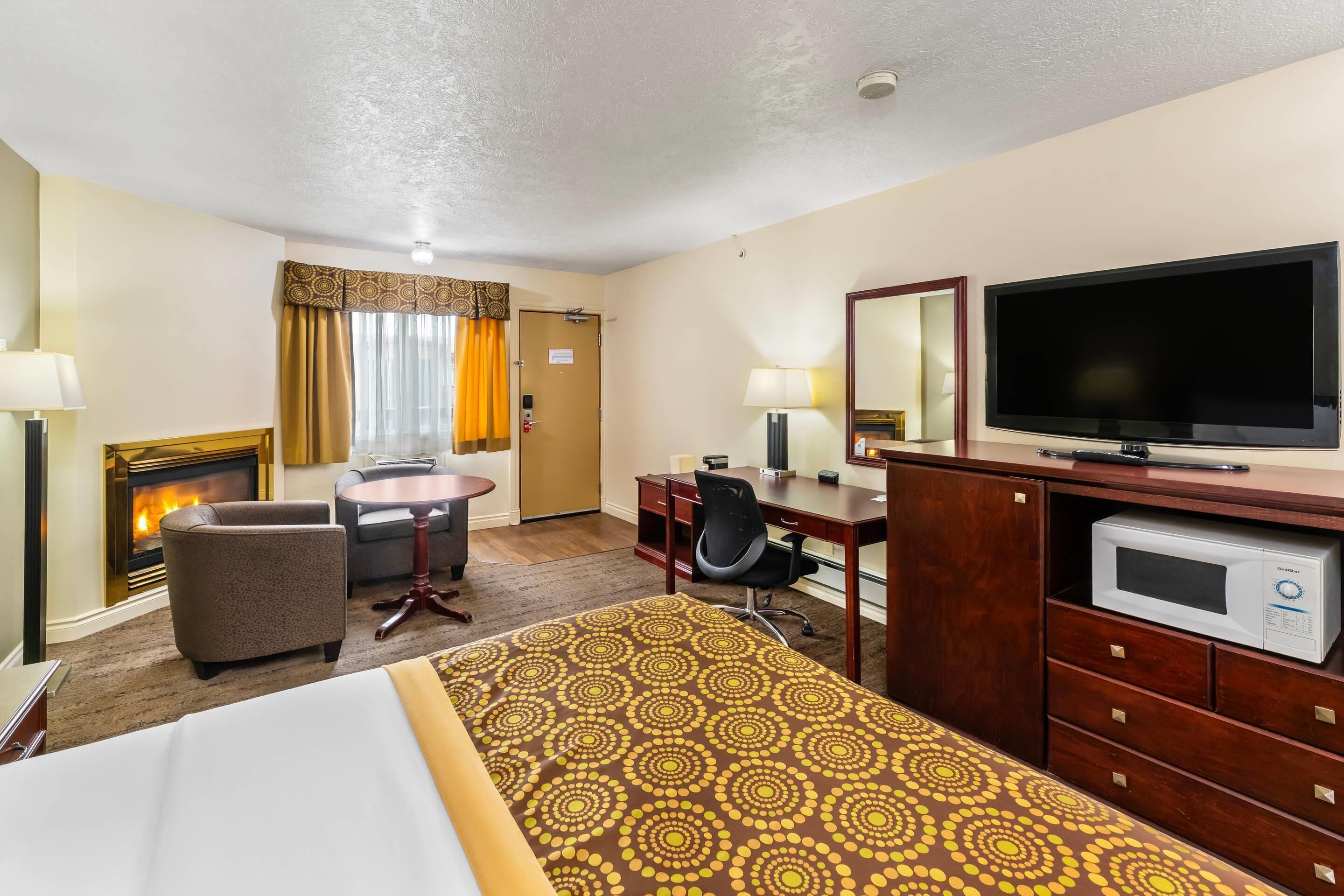 A hotel room with a bed, a desk, a chair, a television, a microwave, and a fireplace.