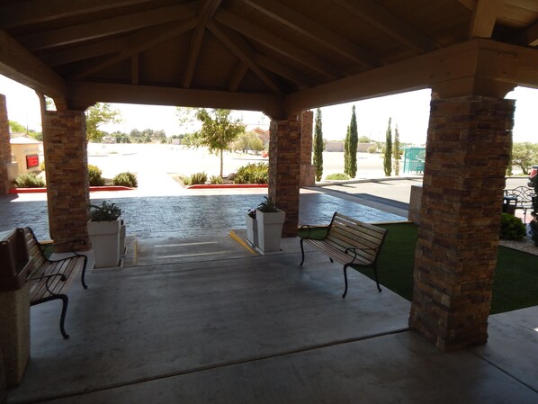 A covered outdoor area with benches and planters, a paved entrance, and a view of a parking lot and buildings in the background.