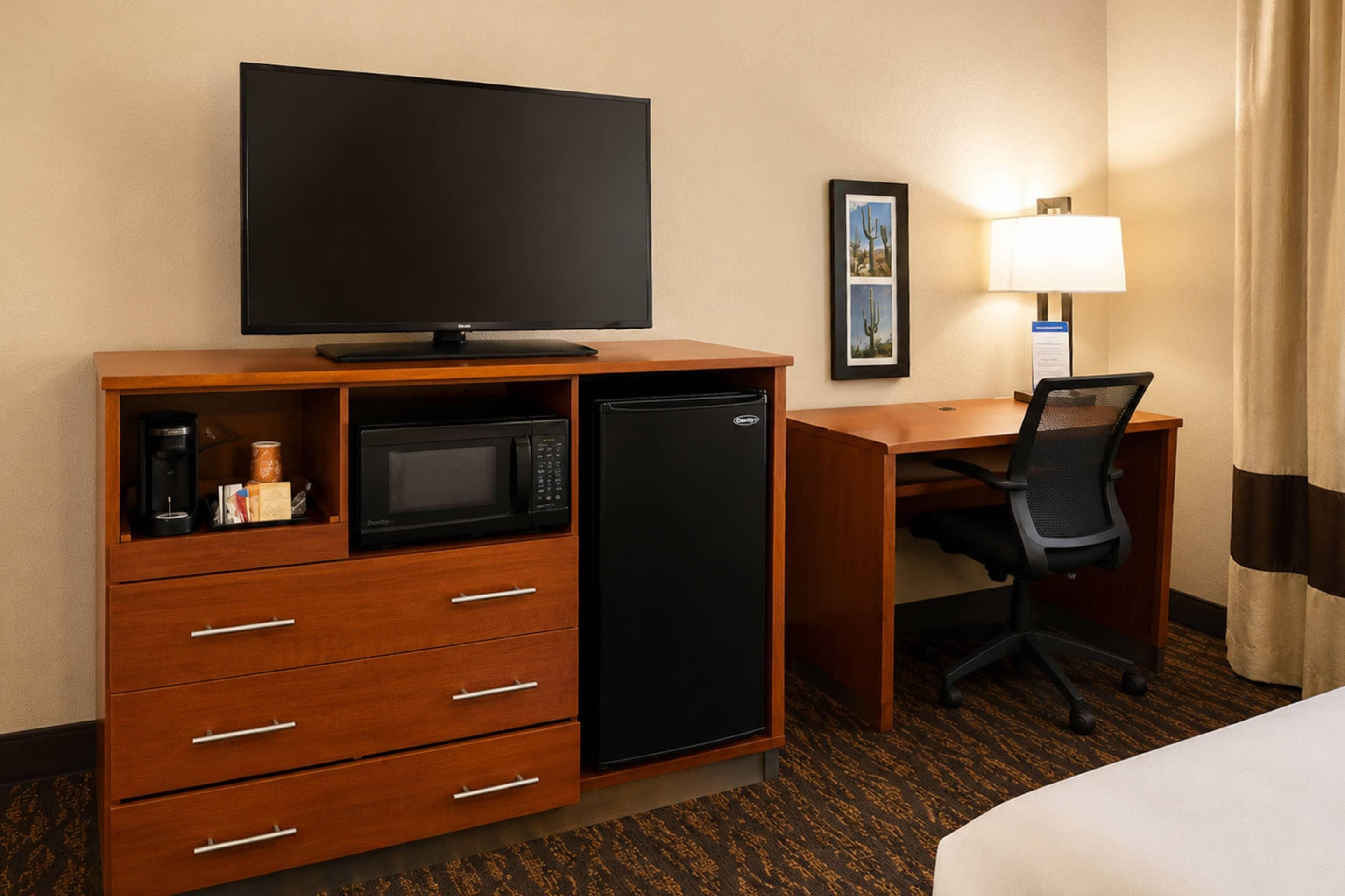 Habitación de hotel con televisión de pantalla plana, microondas, refrigerador, escritorio con silla, lámpara y cama.