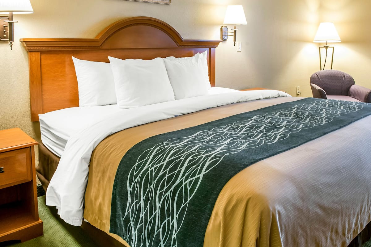 A hotel room with a large bed, wooden headboard, two bedside lamps, a nightstand, and a chair.
