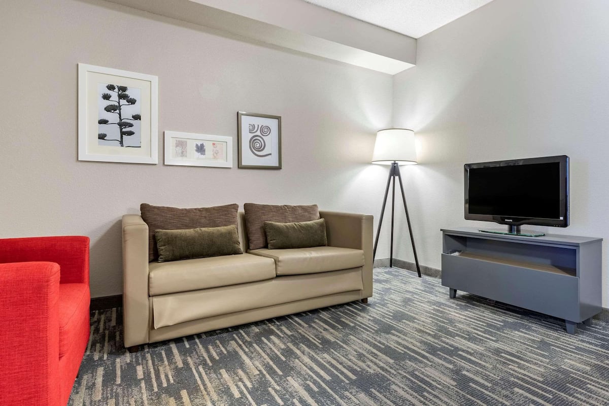 A hotel room with a beige sofa, a red armchair, a flat-screen TV on a grey stand, a floor lamp, and framed artwork on the wall.