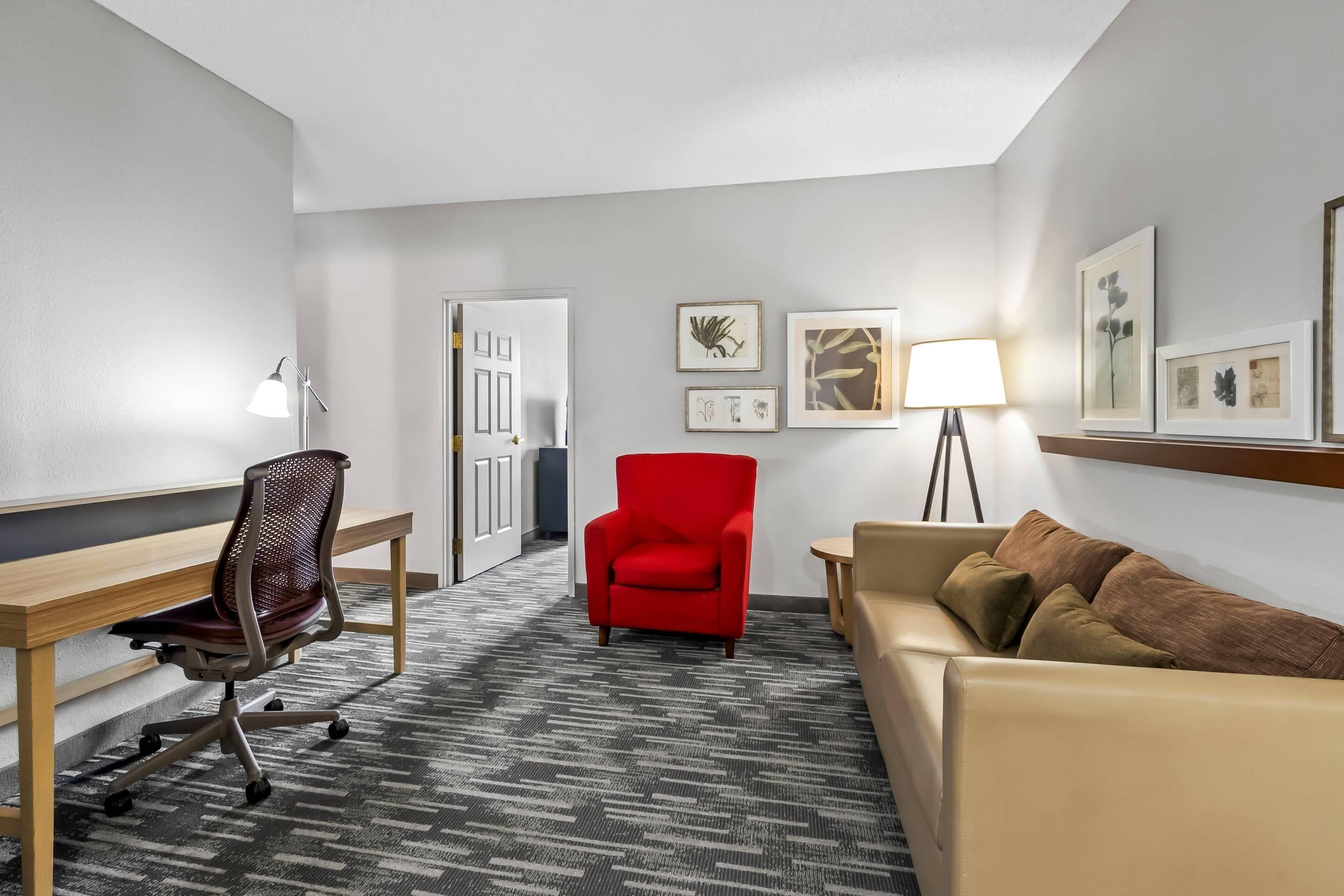 A hotel room with a desk, a red chair, a beige sofa, a patterned carpet, and framed artwork on the walls.