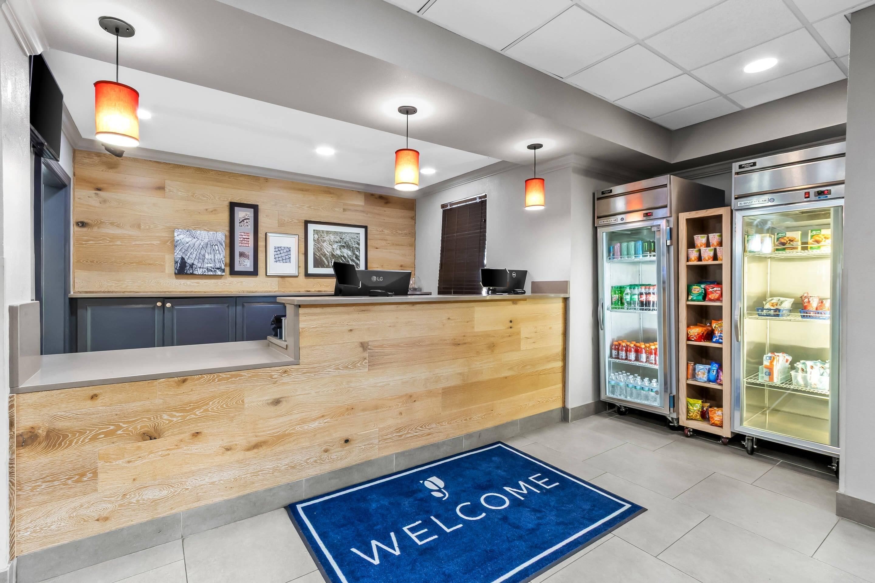A reception area with a wooden counter, two LG monitors, a welcome mat, and a refrigerator stocked with beverages.