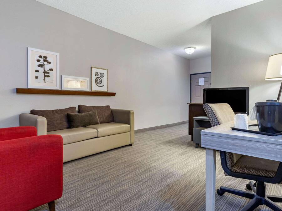A hotel room with a sofa, a red chair, a desk with a lamp, and artwork on the wall.