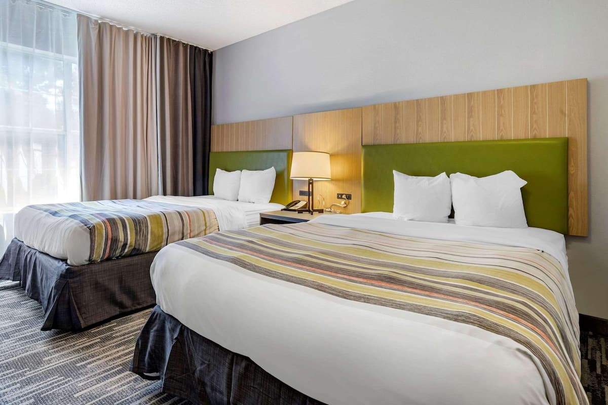 A hotel room with two beds, a large window with curtains, a wooden headboard, and a bedside table with a lamp.