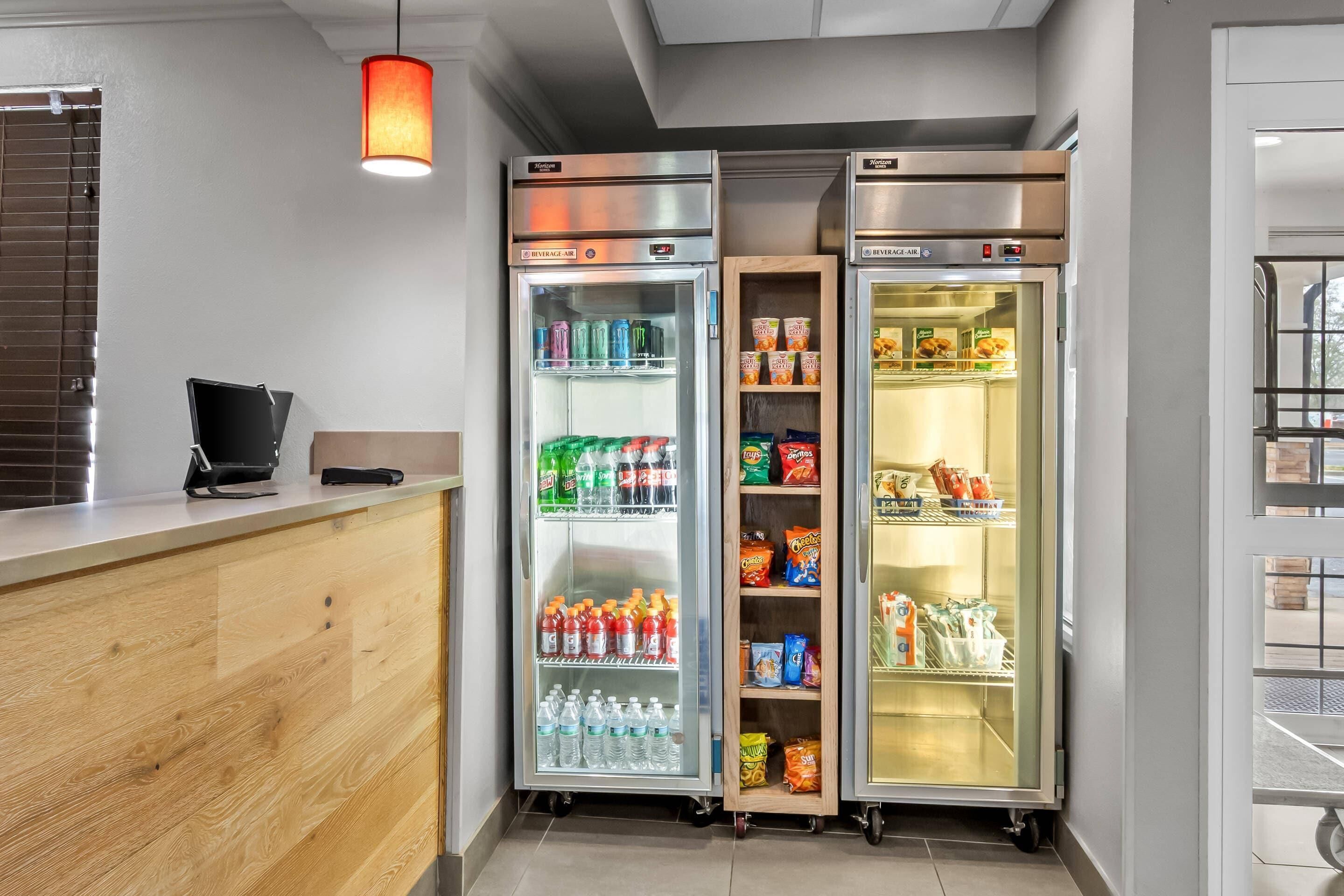 A hotel reception area with a wooden counter, two beverage refrigerators stocked with drinks, and a vending machine.