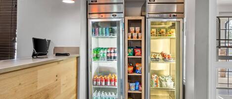 A hotel reception area with a wooden counter, two beverage refrigerators stocked with drinks, and a vending machine.
