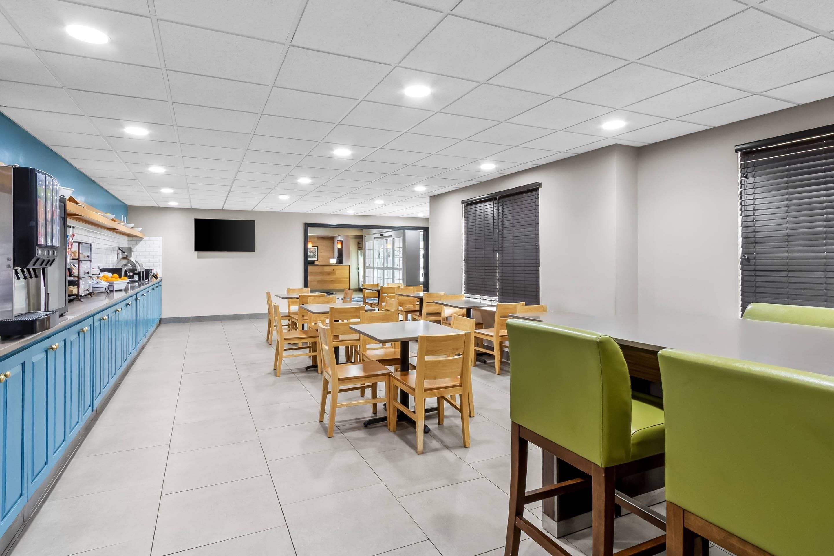 A clean, well-lit dining area with wooden tables and chairs, a blue counter with kitchen appliances, and a large screen on the wall.