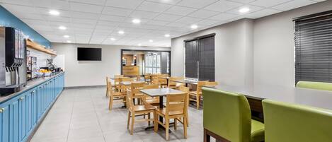 A clean, well-lit dining area with wooden tables and chairs, a blue counter with kitchen appliances, and a large screen on the wall.