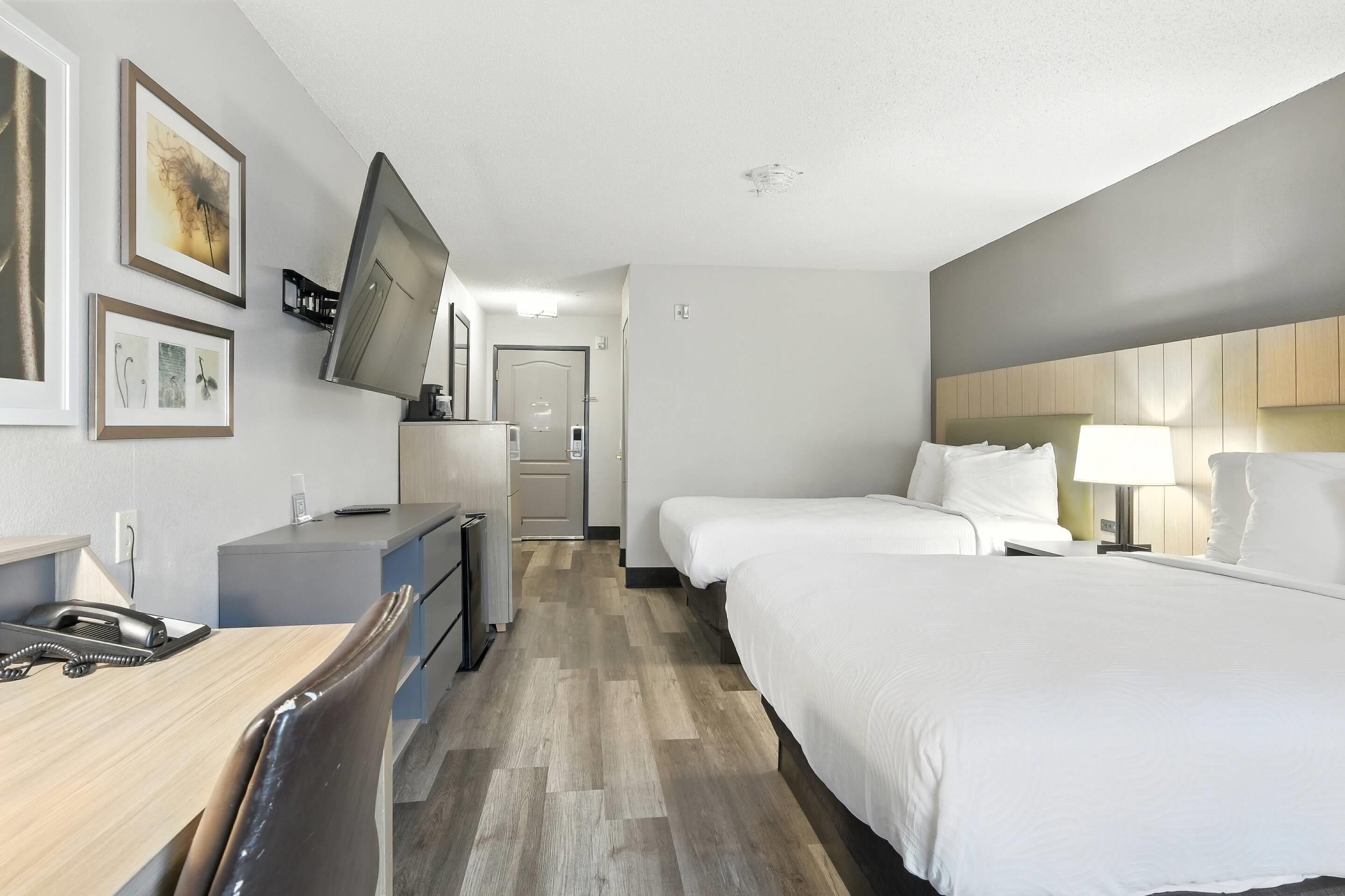 A hotel room with a desk, two beds, a TV mounted on the wall, and a kitchenette.