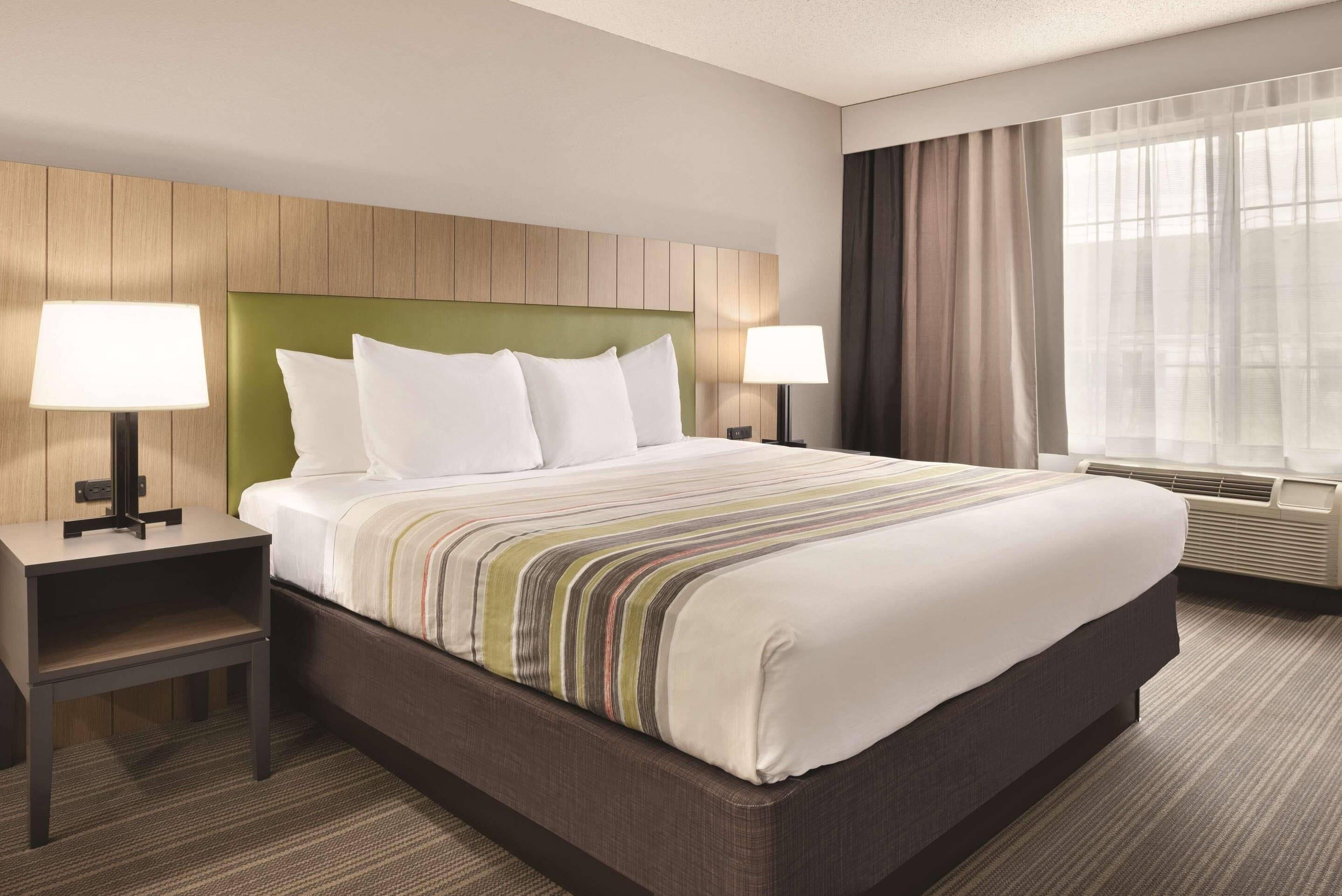 A hotel room with a large bed, two bedside tables with lamps, a green headboard, and a window with curtains.