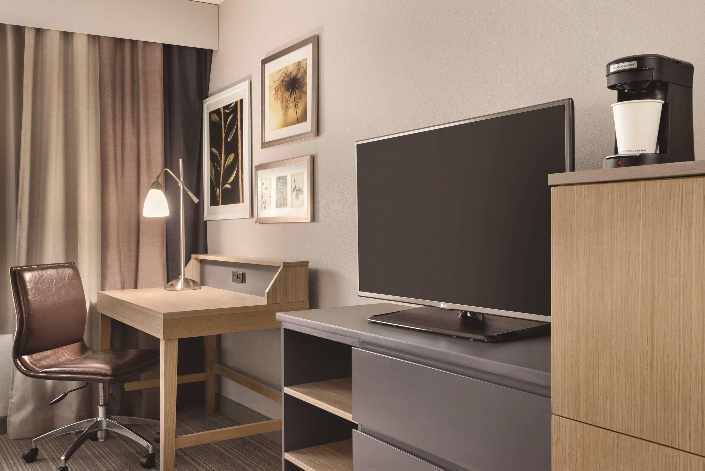 A hotel room with a desk, chair, television, and a coffee machine.