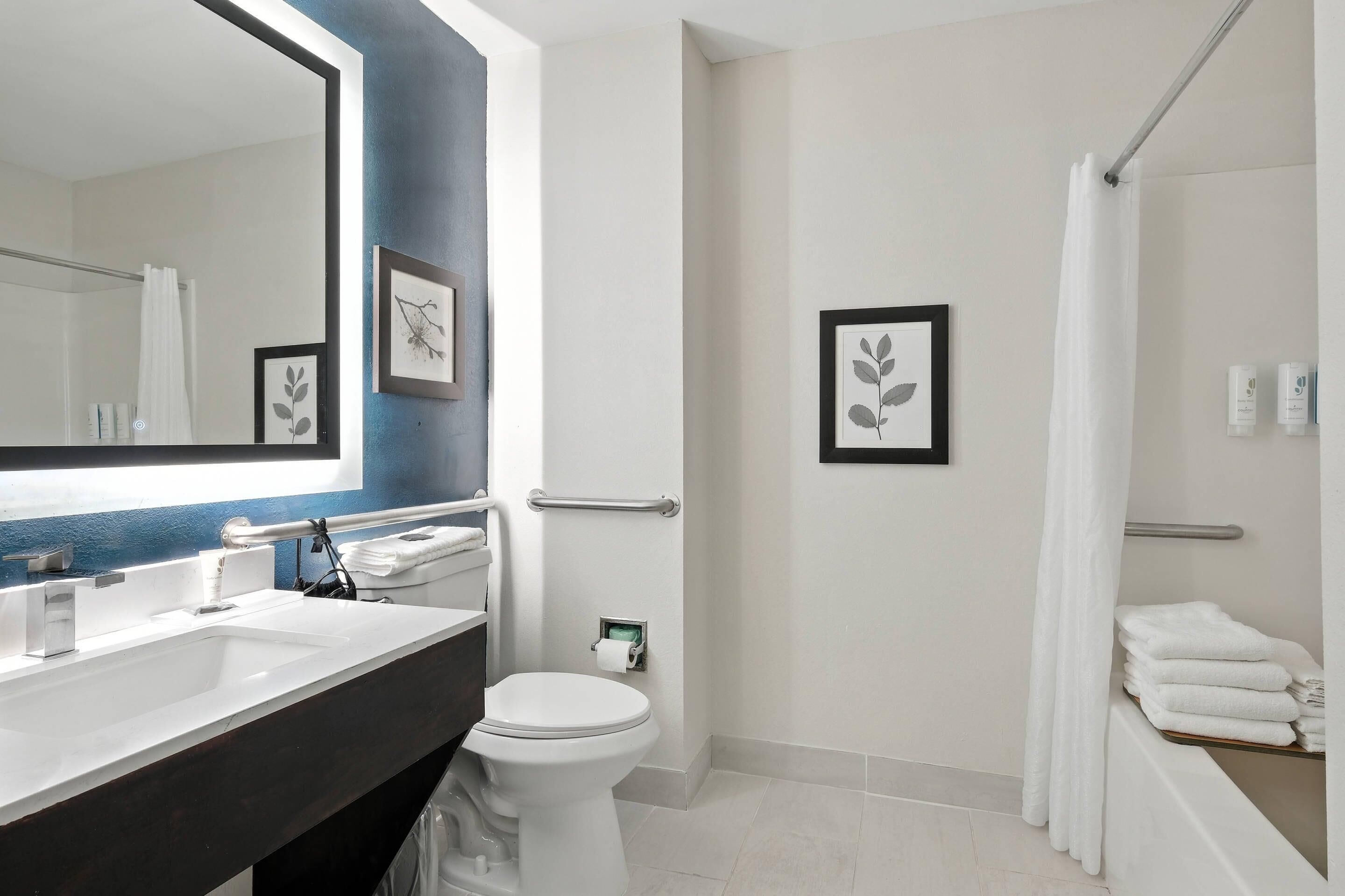 A modern bathroom with a large mirror, white fixtures, a towel rack, and framed artwork on the wall.