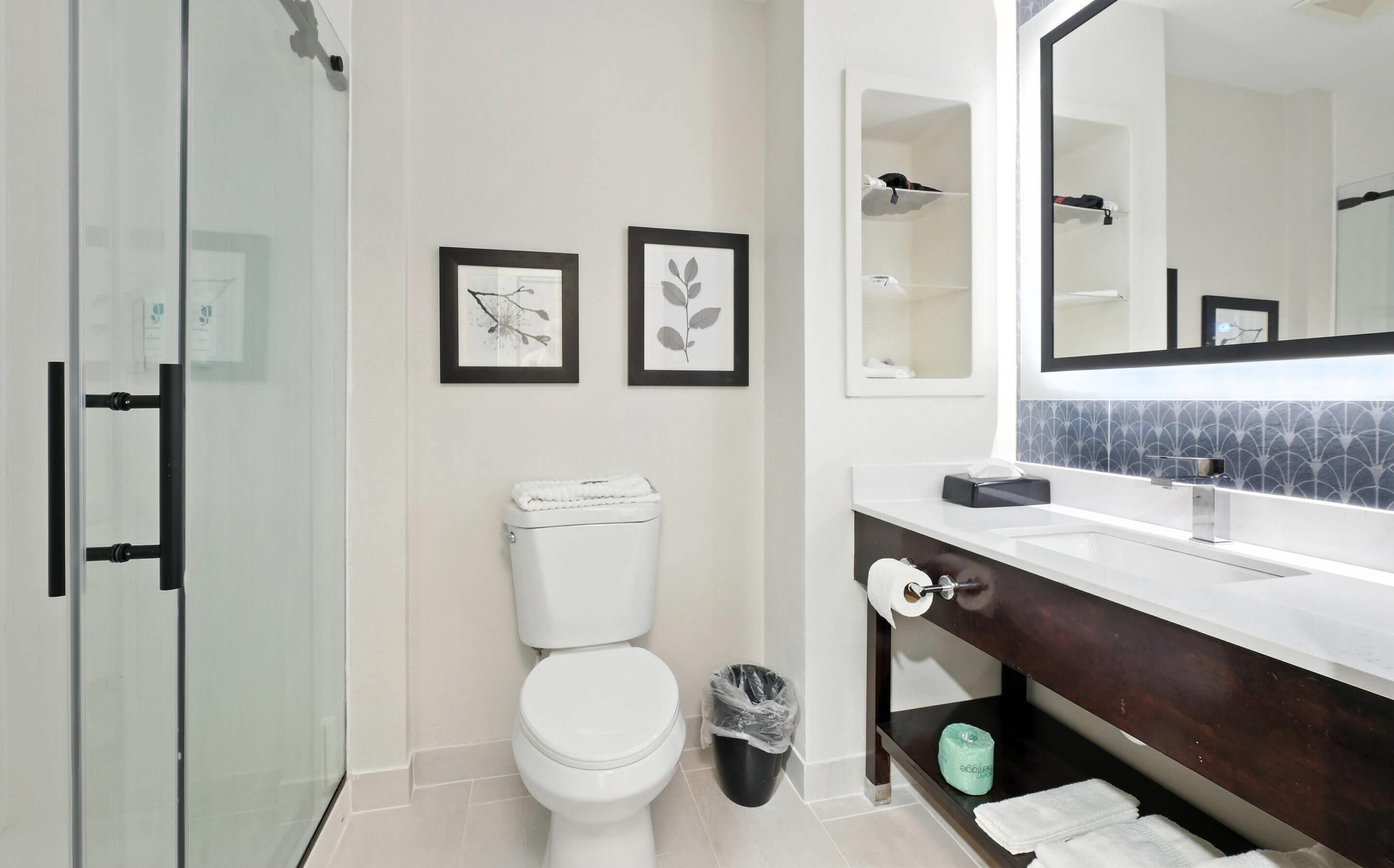 Un baño moderno con inodoro, cabina de ducha de vidrio, lavabo con tocador oscuro y dos cuadros en la pared.