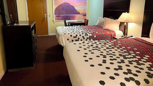 A hotel room with two beds, a desk, a chair, a TV, and a painting of a mountain landscape. - Morro Bay Beach Inn (Morro Bay)