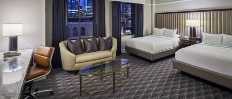A hotel room with a bed, a sofa, a desk with a chair, and a glass coffee table.