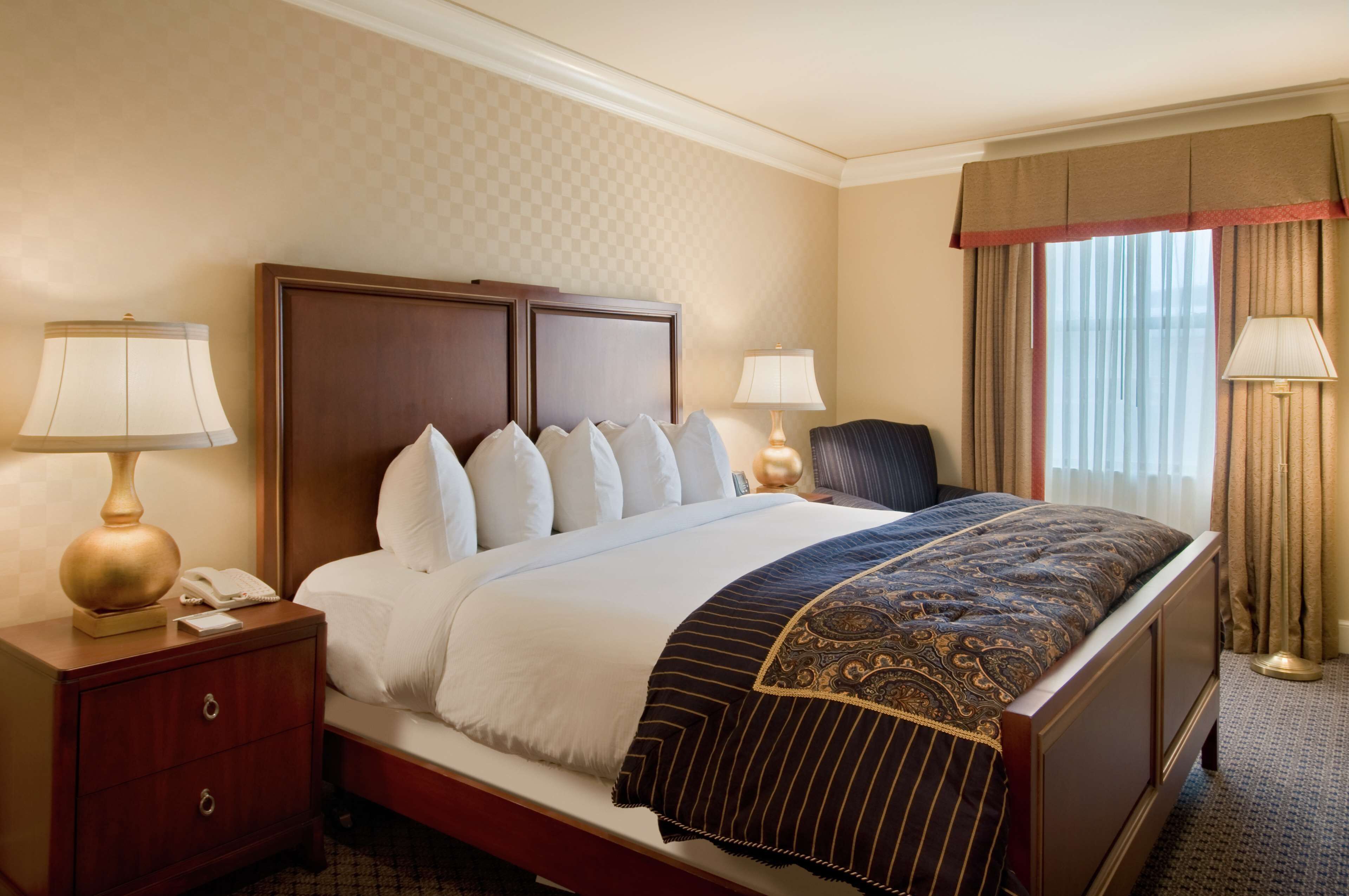 A hotel room with a large bed, two bedside tables with lamps, a nightstand, and a window with curtains.