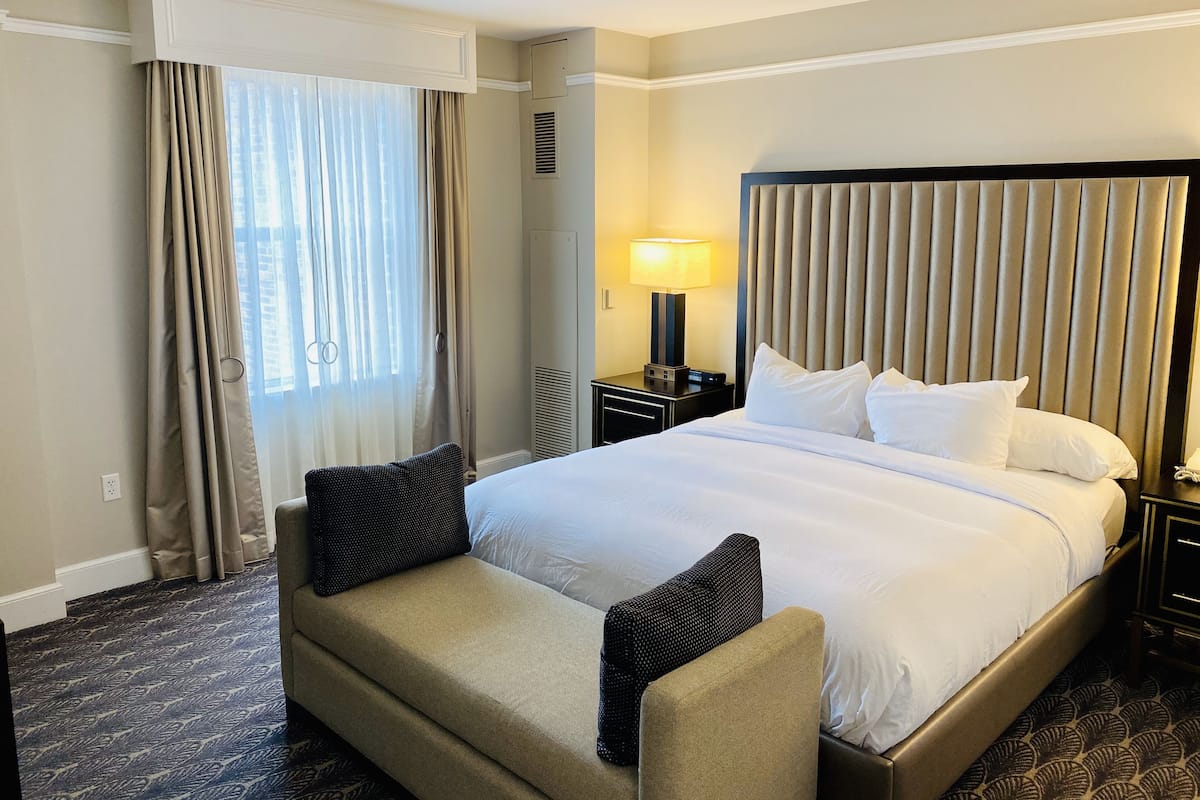 A hotel room with a large bed, a bedside table with a lamp, a window with curtains, and a patterned carpet.