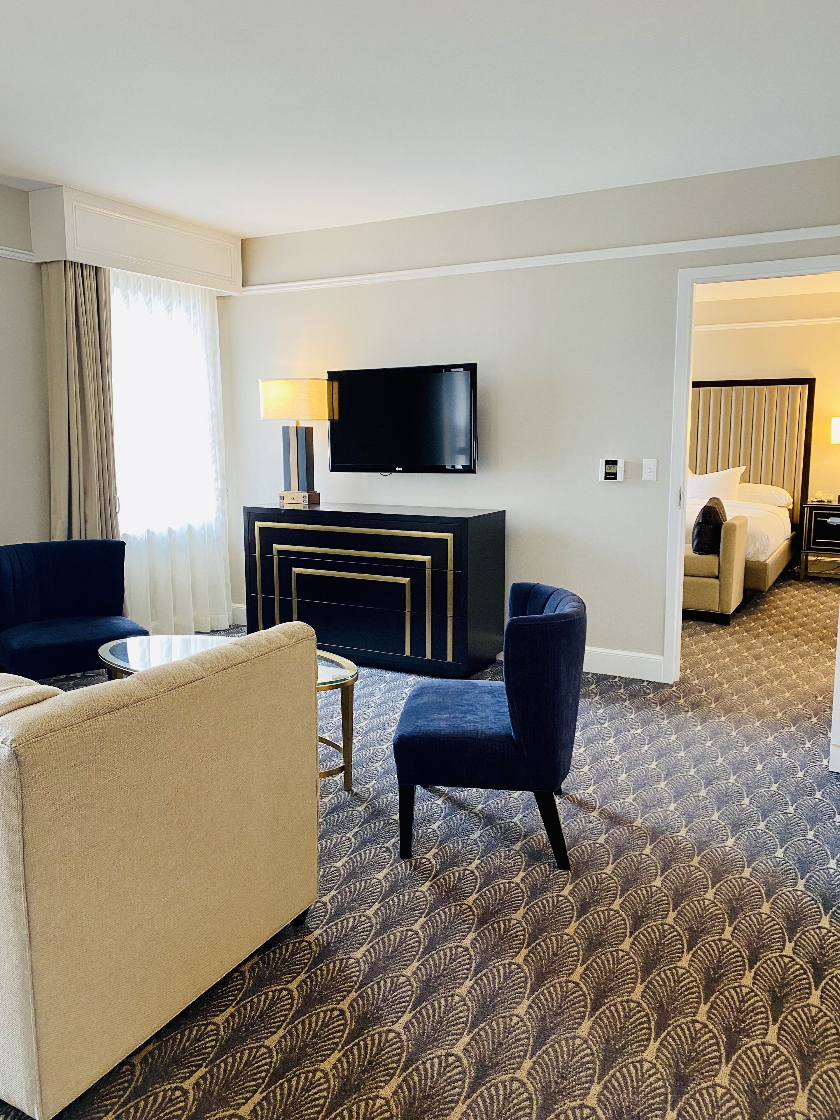 Ein Hotelzimmer mit flachbildfernseher an der Wand, einer Couch, einem Sessel, einem kleinen Tisch und einem gemusterten Teppich.