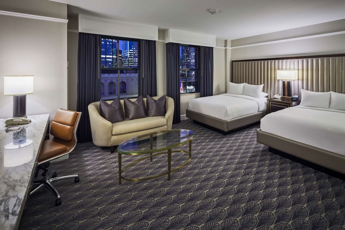 A hotel room with a bed, a sofa, a desk with a chair, a glass coffee table, and a view of the cityscape.