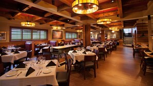 Un restaurant doté de nombreuses tables, recouvertes de nappes blanches, et de chaises, avec des poutres en bois au plafond et de grandes fenêtres équipées de stores.
