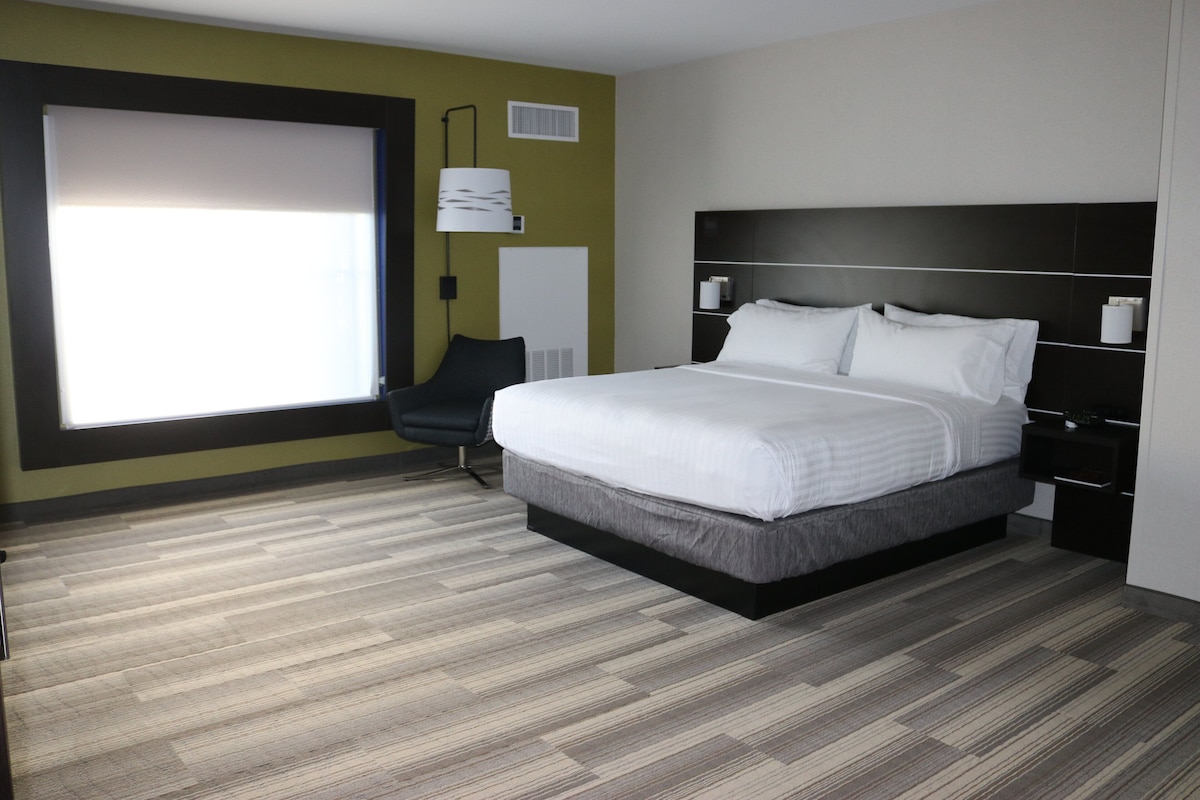 A modern hotel room with a large bed, a chair, a bedside table, and a window.