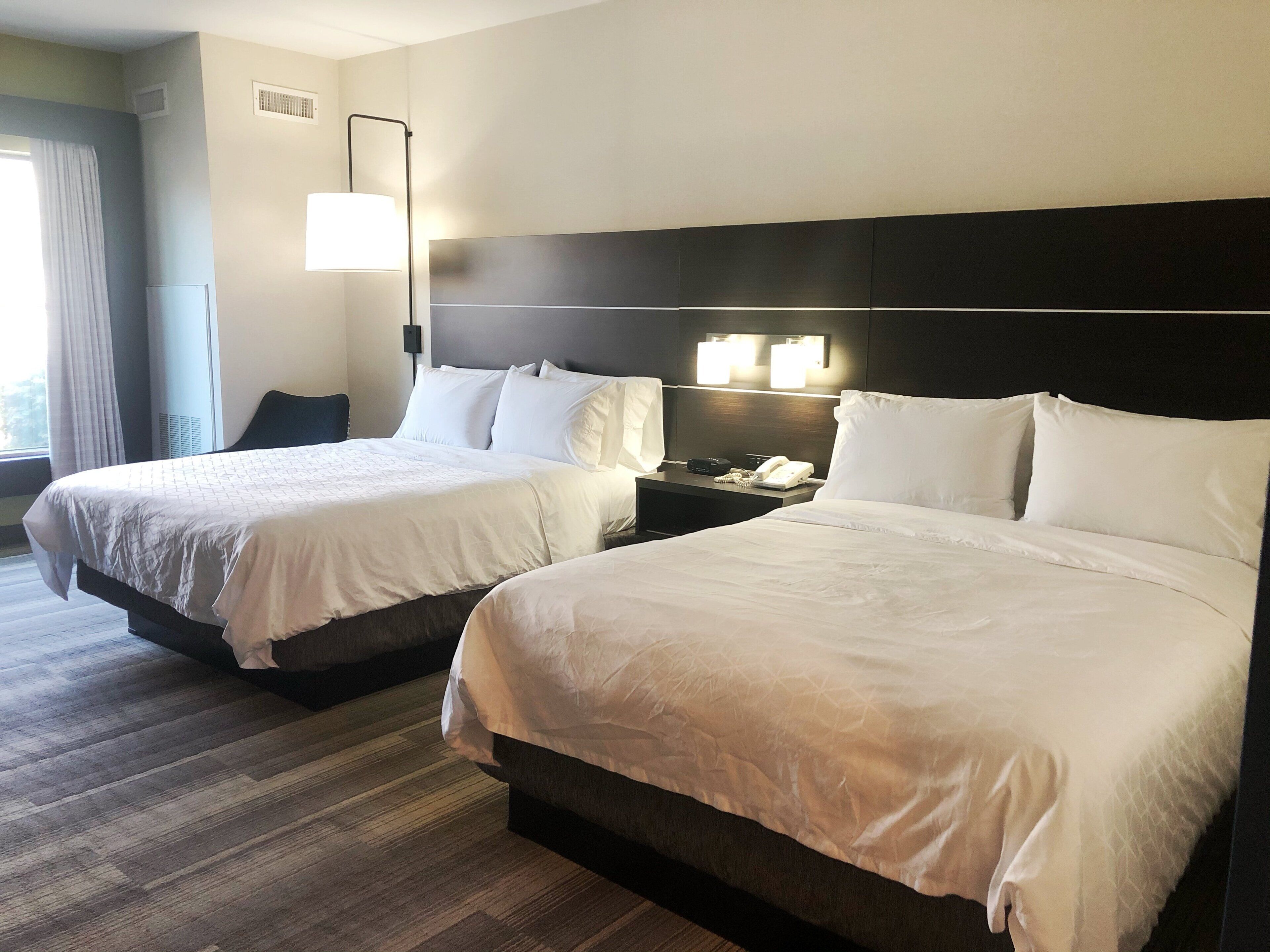 Habitación de hotel con dos camas, una mesita de noche, una lámpara y un ventanal con cortinas.