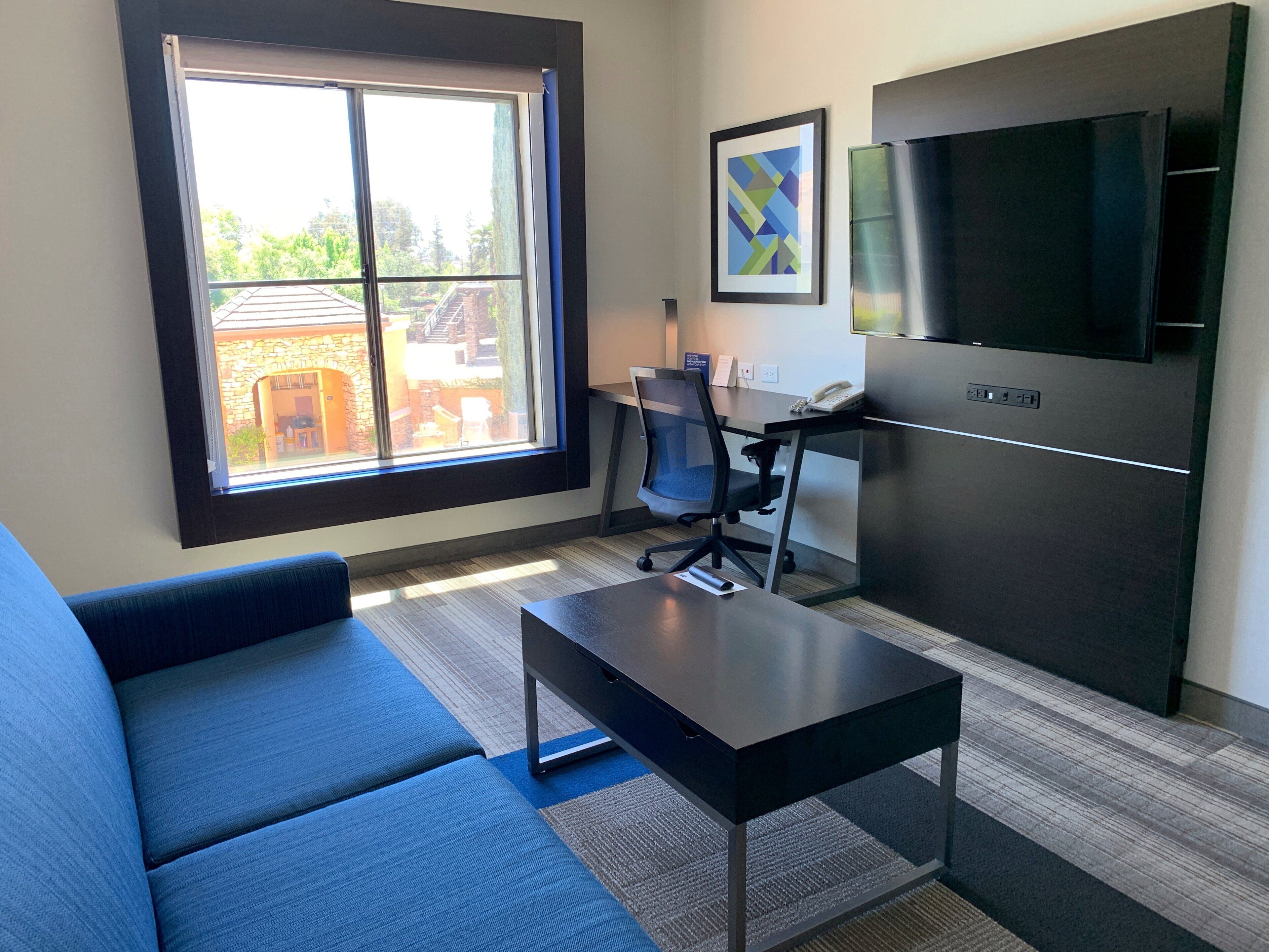 A modern living room with a sofa, a coffee table, a desk with a chair, a flat-screen TV, and a window with a view of a building and trees.