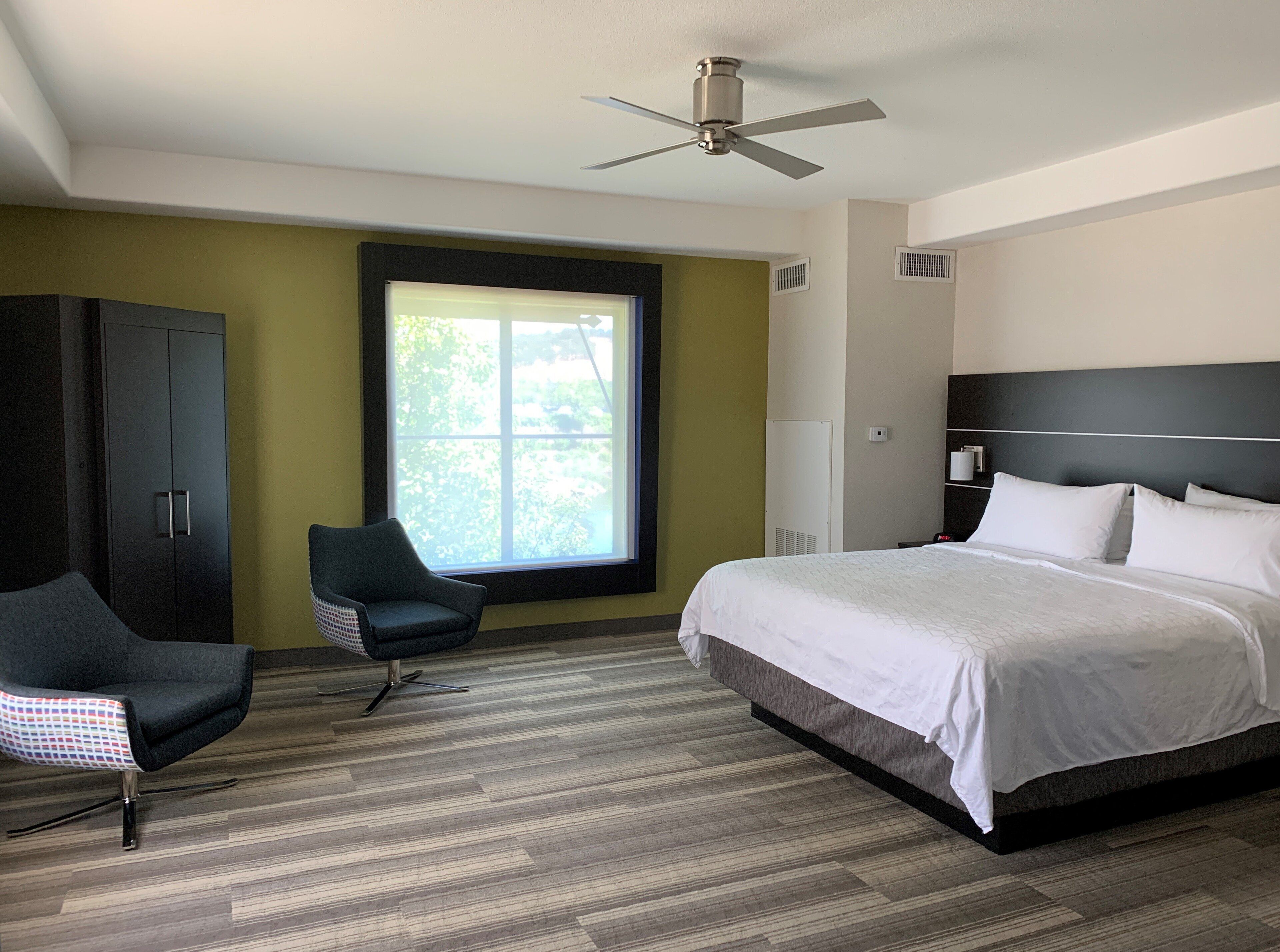A hotel room with a bed, a chair, a wardrobe, a window, and a ceiling fan.