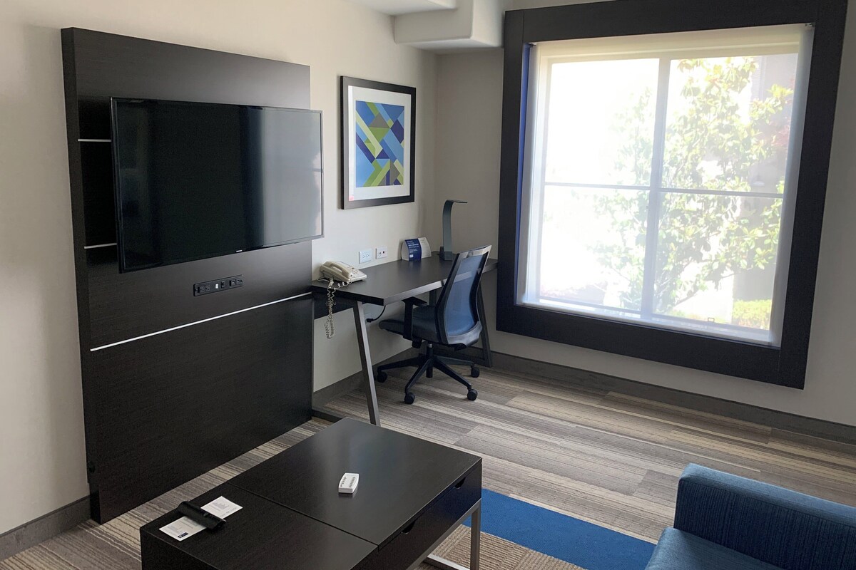Un espacio de oficina moderno con una televisión de pantalla plana grande, un escritorio con una silla, un sofá y un amplio ventanal con vistas a zonas verdes.