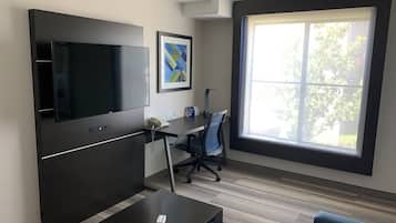 A modern office space with a large flat-screen TV, a desk with a chair, a sofa, and a large window offering a view of greenery.