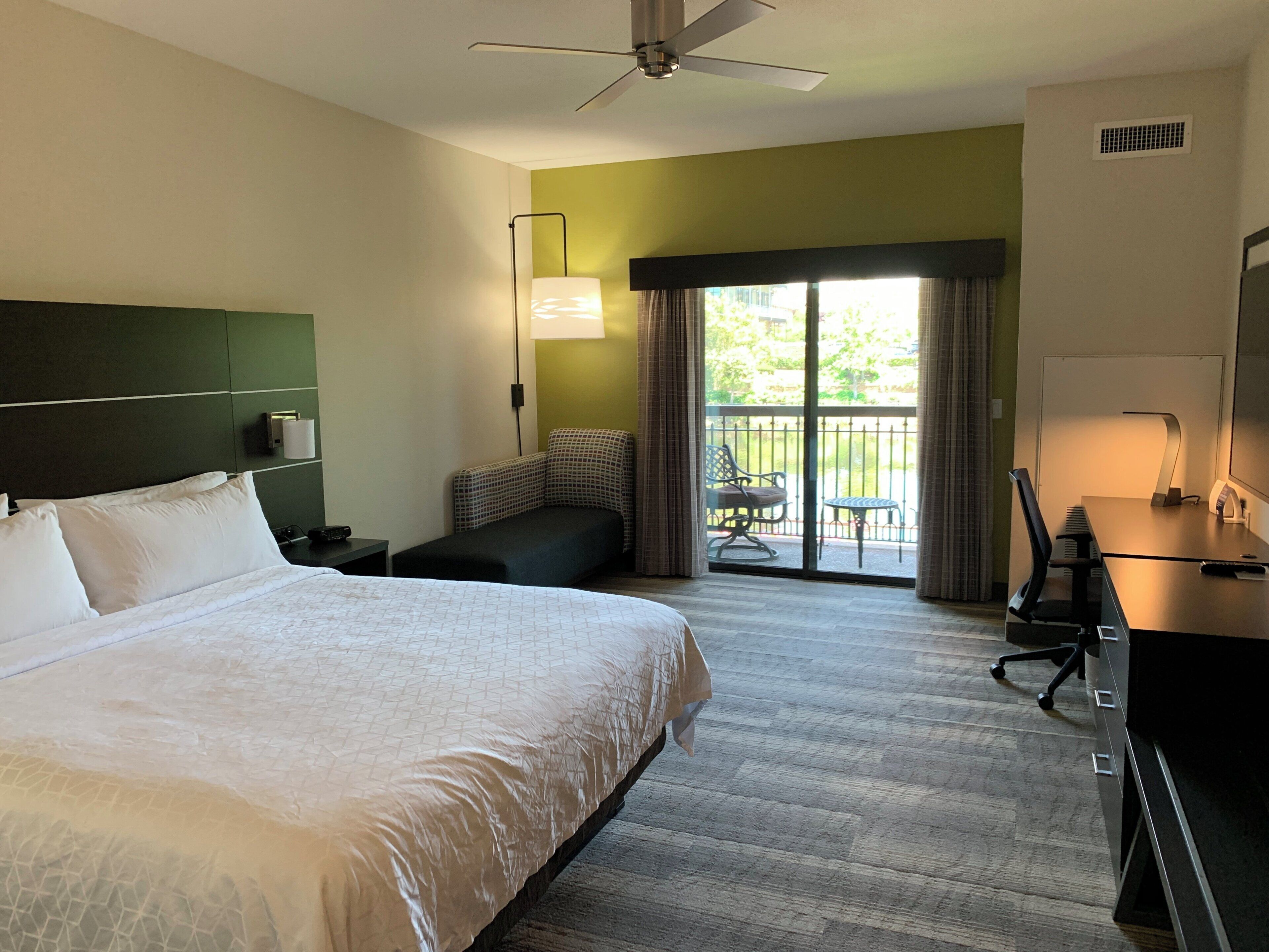 A hotel room with a large bed, a desk with a chair, a balcony with a table and chairs, and a ceiling fan.