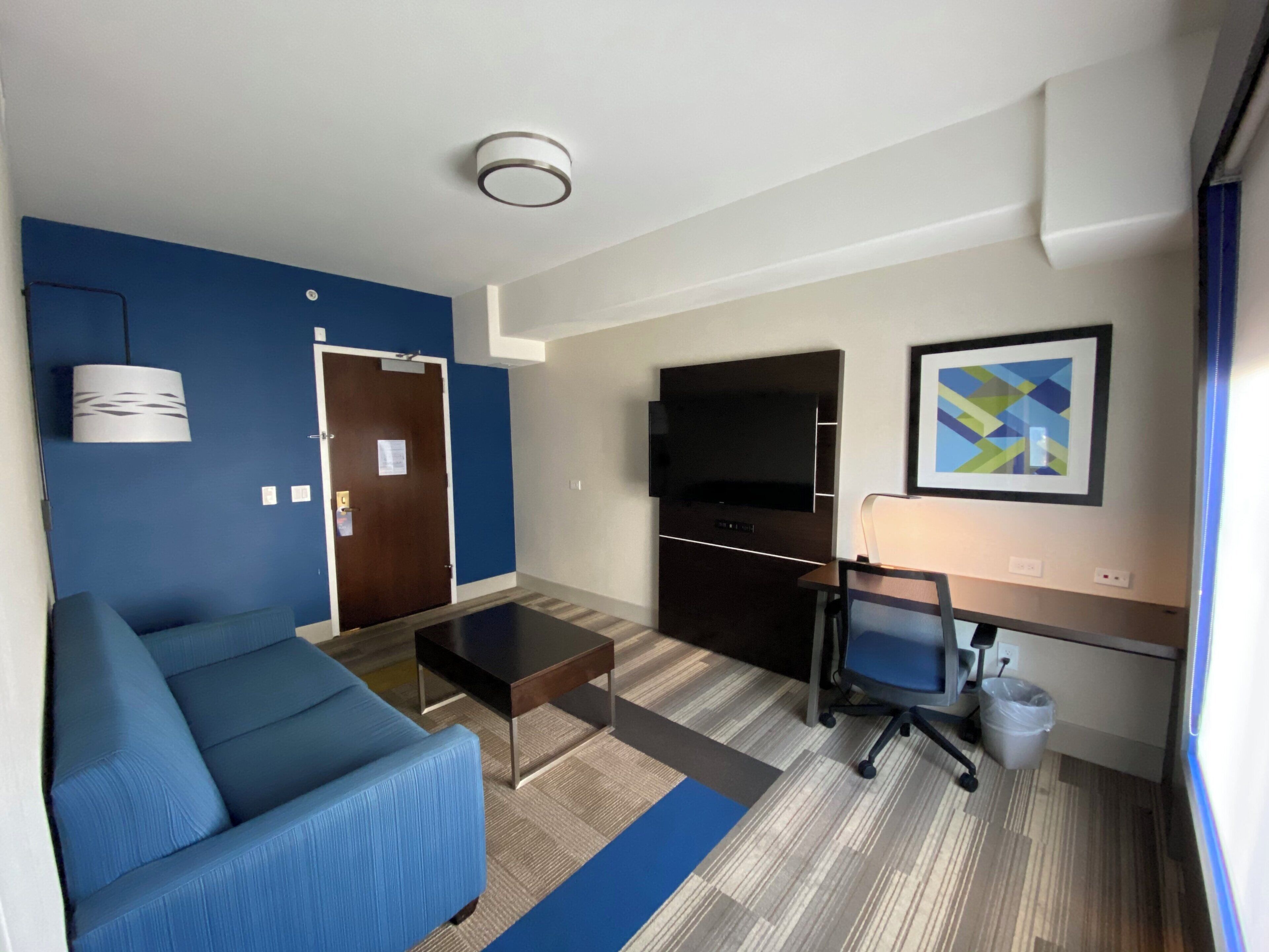 Une chambre d’hôtel moderne avec un canapé bleu, un bureau avec un ordinateur, un téléviseur à écran plat et un petit tableau accroché au mur.