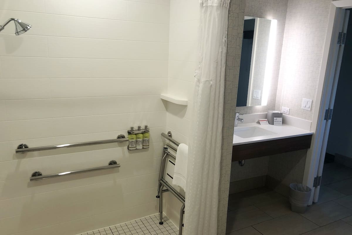 Une salle de bain avec des murs recouverts de carreaux blancs, une douche équipée d’un pommeau de douche à main, d’une barre d’appui et d’un lavabo blanc avec un miroir au-dessus.
