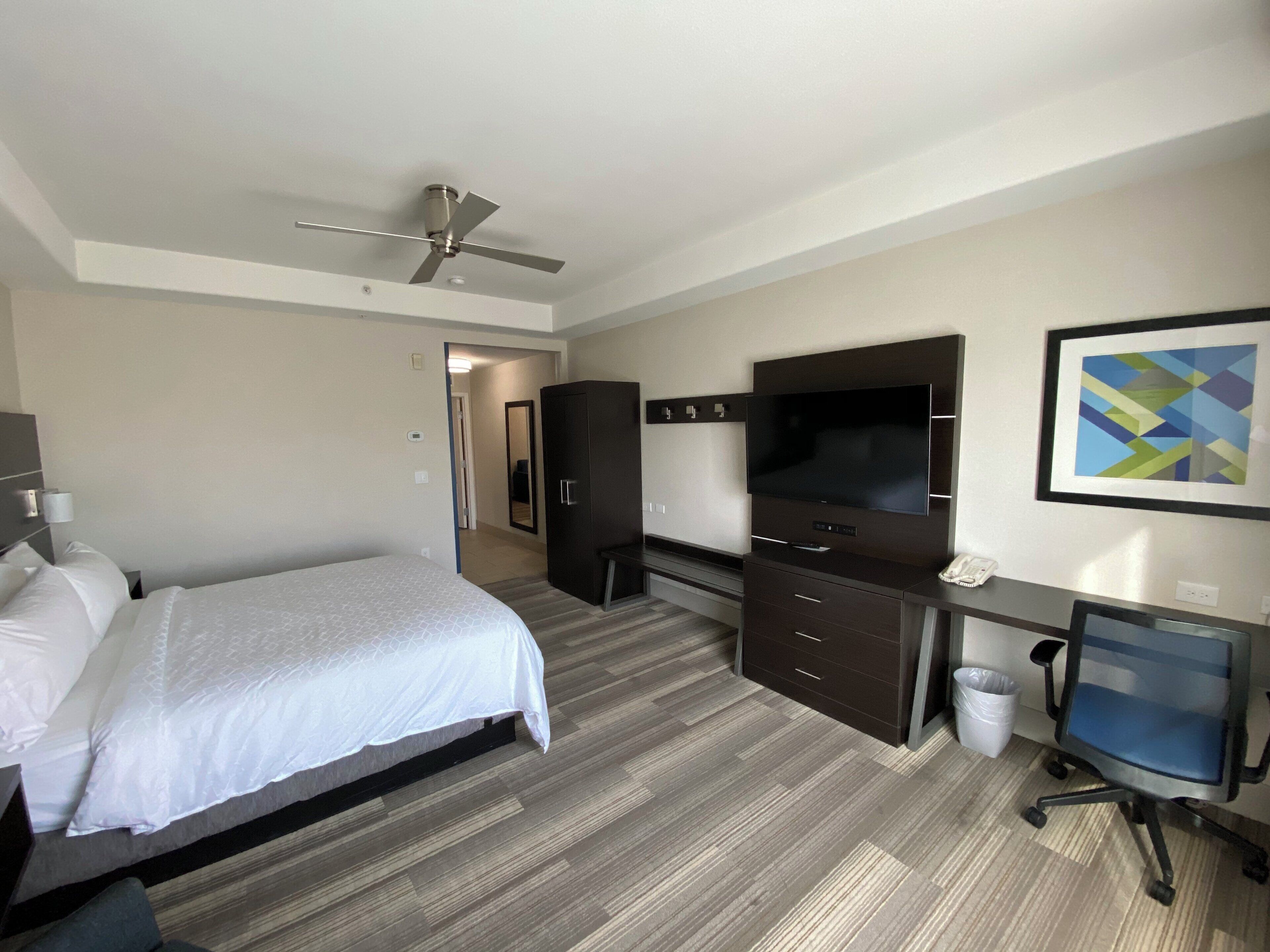 Une chambre d’hôtel équipée d’un lit, d’un bureau avec un ordinateur, d’une télévision, d’une armoire et d’un ventilateur de plafond.