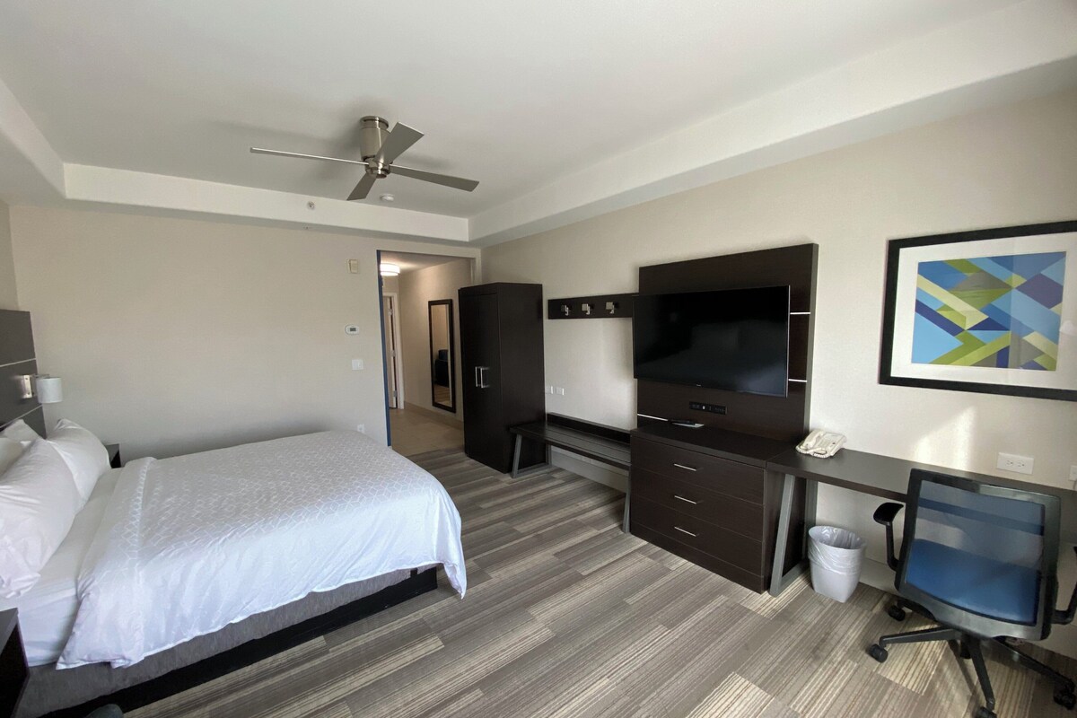 Een hotelkamer met een bed, een bureau met een computer, een televisie, een kledingkast en een plafondventilator.