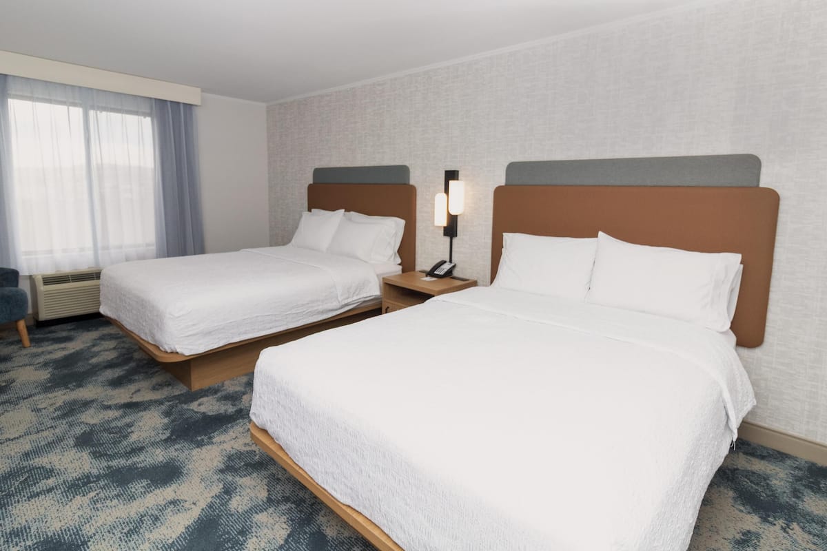 A hotel room with two beds, a nightstand with a lamp, a window with curtains, and a patterned carpet.