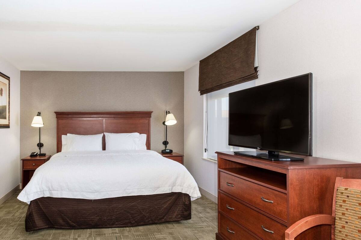 A hotel room with a bed, bedside lamps, a TV, a wooden dresser, and a window with a blind.